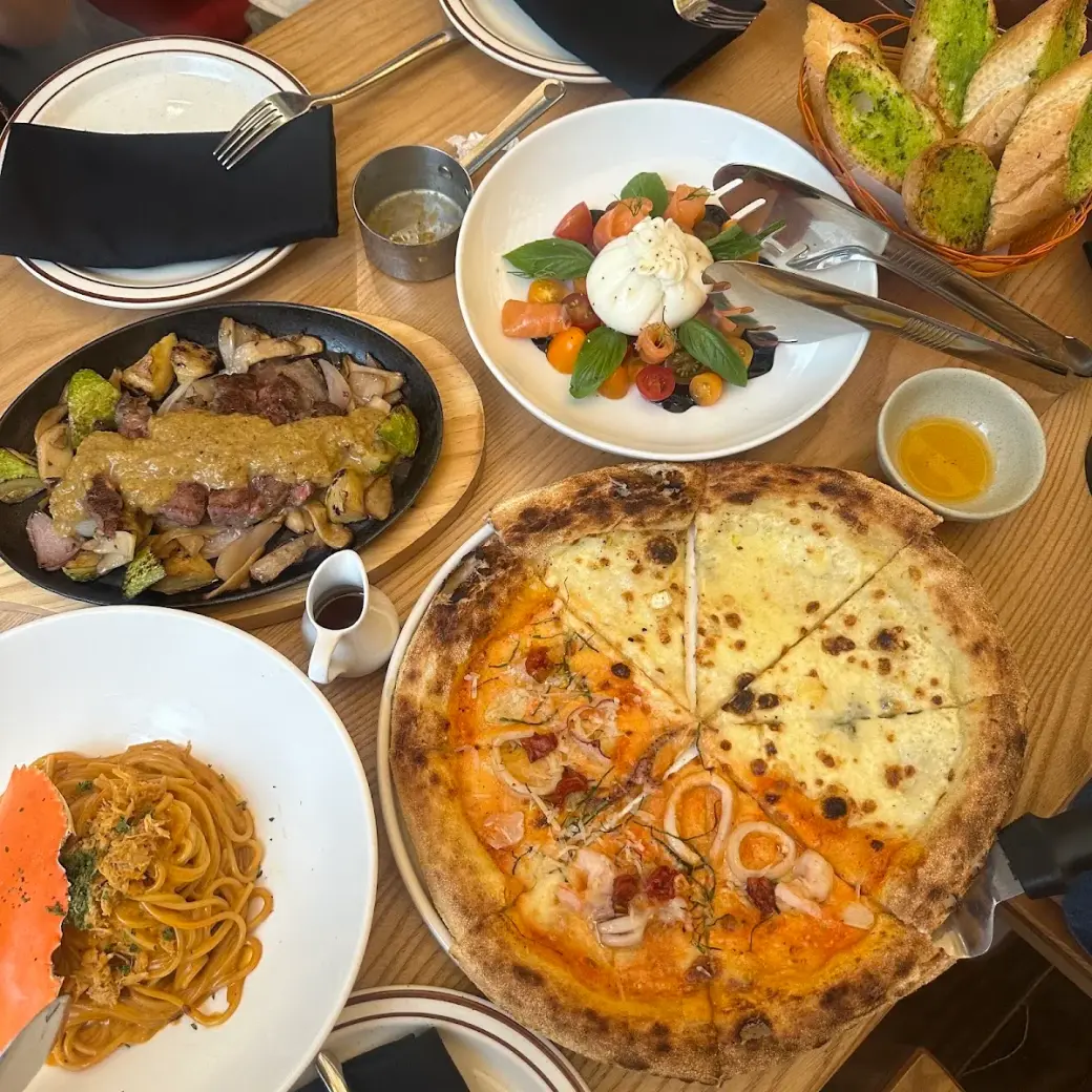 Mâm ăn nhìn từ trên: pizza half-half, burrata, bò xèo, mì Ý và bánh mì bơ tỏi pesto trên bàn gỗ sồi